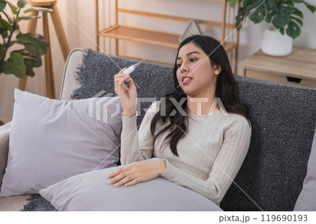 Young Woman Feeling Unwell on Sofa Checking Temperature with Thermometer in Cozy Home Environment 119690193