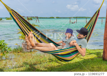 Father with his toddler son lying in a hammock against the backdrop of turquoise waters of Bacalar Lake, Mexico. Peaceful tropical travel destination concept 119691277