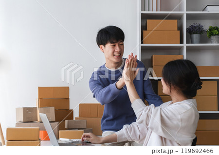 Happy Couple Running a Small Online Business from Home, Surrounded by Cardboard Boxes and Working on a Laptop, Celebrating Success with a High Five in a Bright Modern Workspace Happy Couple Running a Small Online Business from Home, Surrounded by Cardboard Boxes and Working on a Laptop, Celebrating Success with a High Five in a Bright Modern Workspace 119692466