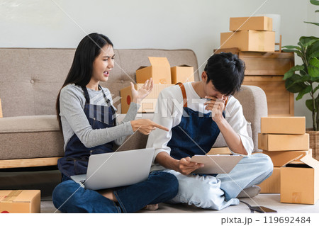 Stressed Couple Managing Online Sales for Small Business at Home with Laptop and Clipboard Surrounded by Cardboard Boxes 119692484