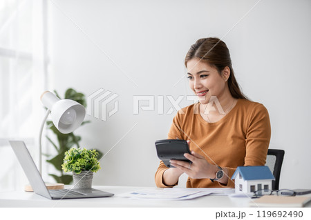 Professional Real Estate Agent Woman Holding House Model and Calculator in Modern Office Setting, Smiling Confidently, Ready to Assist Clients with Property Investments and Home Purchases Professional Real Estate Agent Woman Holding House Model and Calculator in Modern Office Setting, Smiling Confidently, Ready to Assist Clients with Property Investments and Home Purchases 119692490