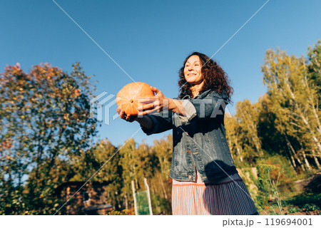Joyful woman tossing a pumpkin on a sunny autumn day in the countryside. Happy woman on her garden plot celebrating autumn and the joy of the harvest 119694001