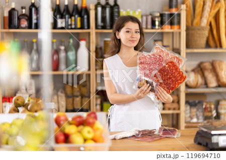 Young saleswoman is offering dried sausages and meat in grocery store Young saleswoman is offering dried sausages and meat in grocery store 119694710