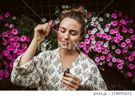 Beautiful woman doing her skin care routine outdoors, wearing bathrobe. Woman using facial skin serum with dropper. 119695021
