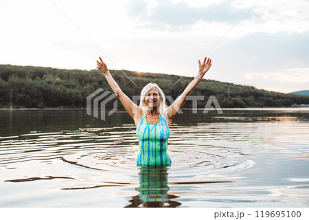 Cold water swimming for elderly women. Senior sporty women standing in lake during cold evening, arms in the air. Cold water swimming for elderly women. Senior sporty women standing in lake during cold evening, arms in the air. 119695100