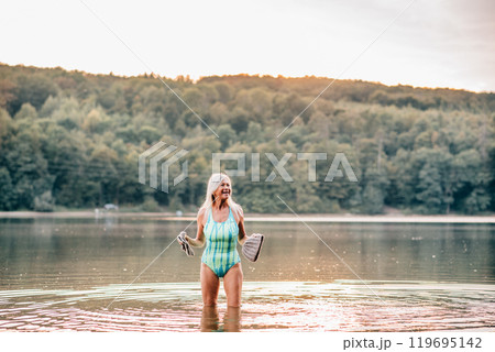 Cold water swimming for elderly women. Portrait of senior sporty women standing in lake during cold evening, holding gloves and hat in hads. 119695142