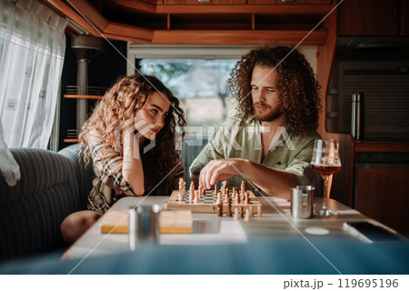 Young couple is on a camping trip in nature, sitting in caravan and playing chess. Caravan traveling for young people. 119695196