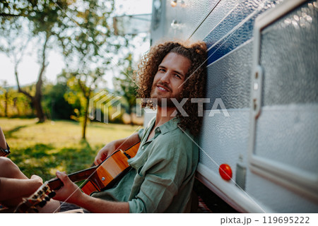 Young man is on a camping trip in nature, sitting in front caravan and playing guitar alone. Young man is on a camping trip in nature, sitting in front caravan and playing guitar alone. 119695222