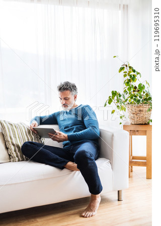 Portrait of attractive middle aged man at home, watching tv show on tablet, sitting on sofa in living room. 119695310