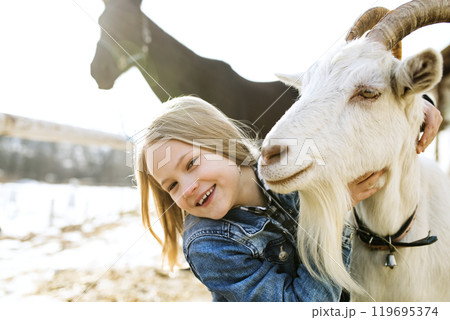 Young girl spending cold winter weekend on a farm, taking care of animals and petting white goat. Young girl spending cold winter weekend on a farm, taking care of animals and petting white goat. 119695374