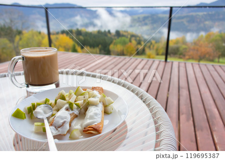 Pancakes with chopped fruits and coffee cup on amazing autumn landscape background. Breakfast in mountains. Breakfast on wooden terrace in mountain resort. Tourism in Ukraine.  119695387