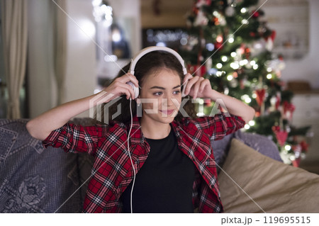 Student having hygge Christmas at home. Woman is sitting on sofa by Christmas tree, listening music with headphones. Student having hygge Christmas at home. Woman is sitting on sofa by Christmas tree, listening music with headphones. 119695515