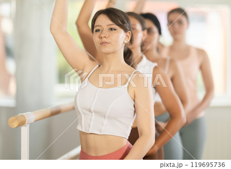 Portrait of pretty young girl practicing 3rd position of ballet stance in classical ballet with group of young beautiful girls in sports clothing in ballet hall Portrait of pretty young girl practicing 3rd position of ballet stance in classical ballet with group of young beautiful girls in sports clothing in ballet hall 119695736