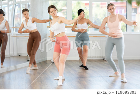 Young female ballet dancer executing dance moves with focused precision among group of people rehearsing in ballet studio 119695803