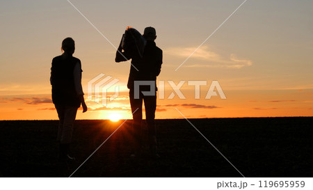 Two farmers are standing in a field at sunset. A man holds a bag on his shoulder, next to his wife Two farmers are standing in a field at sunset. A man holds a bag on his shoulder, next to his wife 119695959