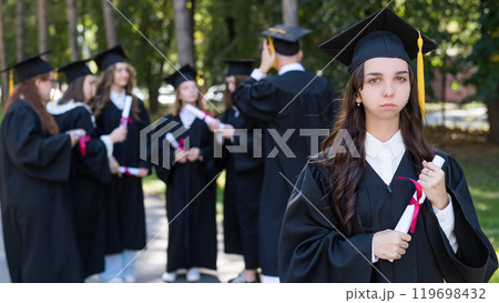 Group of happy students in graduation gowns outdoors. A young girl is holding a diploma. 119698432