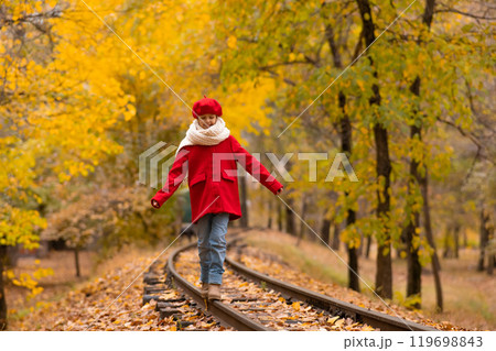 Caucasian girl in a red coat and beret walks along the railway tracks in the park in autumn. Caucasian girl in a red coat and beret walks along the railway tracks in the park in autumn. 119698843