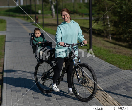 Caucasian woman riding bicycle with toddler son sitting behind her in child seat in park.  119699286