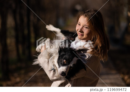 Caucasian woman holding a border collie in her arms while walking in the autumn park. Portrait of a girl with a dog. Caucasian woman holding a border collie in her arms while walking in the autumn park. Portrait of a girl with a dog. 119699472