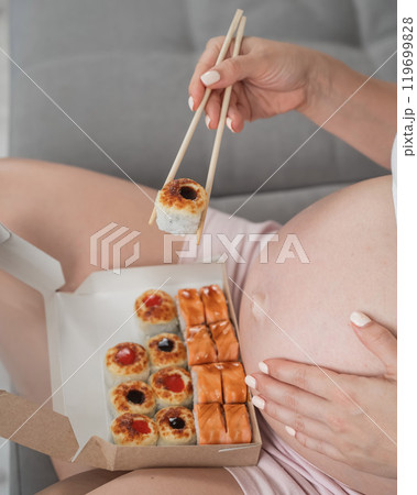 A pregnant woman sits on the sofa and eats rolls from a box. Food delivery. Close up of belly.  119699828