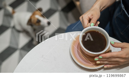Woman drinking coffee in a dog friendly cafe. Jack Russell sits on the floor in a cafe and waits for the owner. Woman drinking coffee in a dog friendly cafe. Jack Russell sits on the floor in a cafe and waits for the owner. 119699867