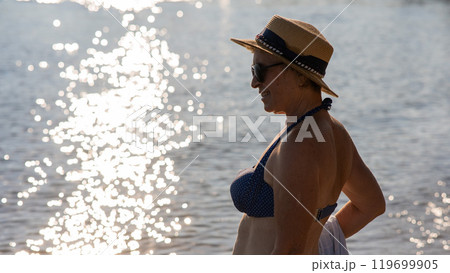 An old woman in a straw hat, sunglasses and a swimsuit is resting on the beach.  119699905
