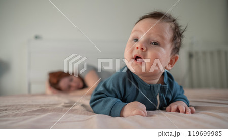 A three-month-old boy lies on his stomach on the bed and his mother sleeps behind him. Postpartum depression. 119699985