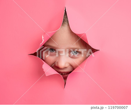 Cute Caucasian girl peeks out of a hole in a paper pink background. Cute Caucasian girl peeks out of a hole in a paper pink background. 119700007
