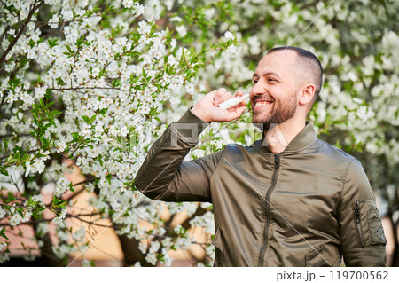 Man allergic using medical nasal drops, suffering from seasonal allergy at spring in blossoming garden. Happy man treating runny nose in front of blooming tree outdoors. Spring allergy concept. 119700562