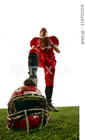 Champion. Bottom view of man, American football player in red uniform, standing with ball, placing leg on helmet lying on stadium. Champion. Bottom view of man, American football player in red uniform, standing with ball, placing leg on helmet lying on stadium. 119703359