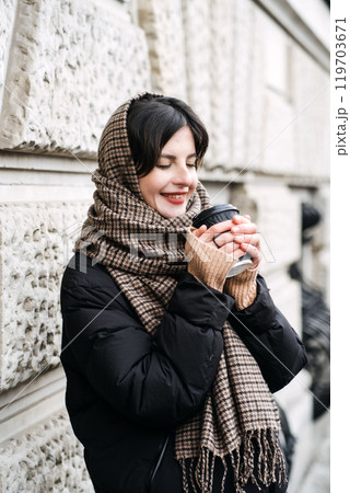 A woman holding a coffee cup wrapped in a warm scarf, combining mindful city living and seasonal comfort essentials. 119703671