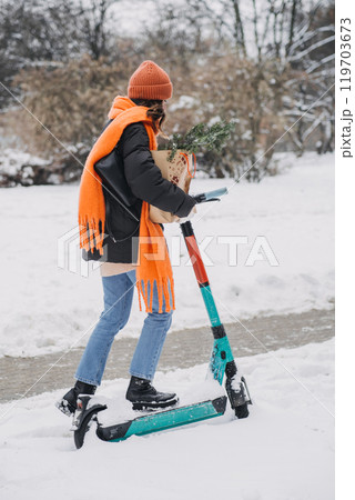 Young woman on e-scooter in winter park wearing orange scarf and beanie, holding gift-wrapped package. Sustainable transport in urban winter holidays, eco-friendly mobility trend 119703673