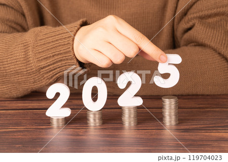 2025 New year saving money and financial planning concept. Female hands putting white number 2025 on stack of coins. Creative idea for business growth, tax payment, investment and banking. 2025 New year saving money and financial planning concept. Female hands putting white number 2025 on stack of coins. Creative idea for business growth, tax payment, investment and banking. 119704023