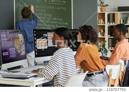 Students working on computers during a lesson with a teacher writing on chalkboard and bookshelf in background. Classroom environment fostering collaborative learning Students working on computers during a lesson with a teacher writing on chalkboard and bookshelf in background. Classroom environment fostering collaborative learning 119707793