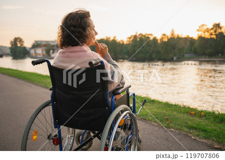 Sad senior woman sitting in wheelchair walking on road in city outdoor, back view. Old paralyzed lady in chair for people with disability feel depressed. Lonely elderly sick woman sits on wheelchair 119707806