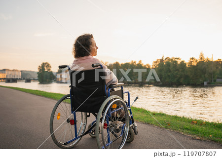 Sad senior woman sitting in wheelchair walking on road in city outdoor, back view. Old paralyzed lady in chair for people with disability feel depressed. Lonely elderly sick woman sits on wheelchair Sad senior woman sitting in wheelchair walking on road in city outdoor, back view. Old paralyzed lady in chair for people with disability feel depressed. Lonely elderly sick woman sits on wheelchair 119707807