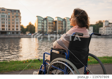 Sad senior woman sitting in wheelchair walking on road in city outdoor, back view. Old paralyzed lady in chair for people with disability feel depressed. Lonely elderly sick woman sits on wheelchair 119707809