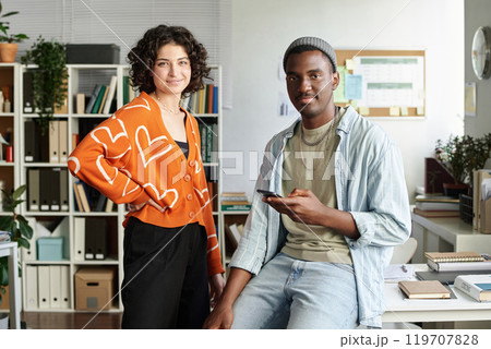 Smiling colleagues collaborating in office setting with shelves and plants in background creating a modern and relaxed atmosphere with gadgets and paperwork on the desk 119707828