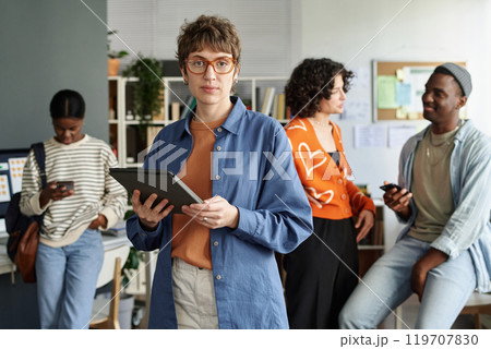 Portrait of woman wearing glasses and blue shirt, holding tablet, standing in modern office with multiple coworkers in background, engaging in various tasks 119707830