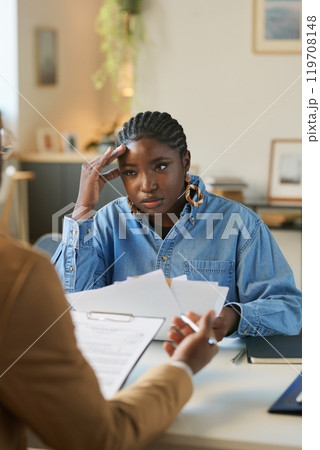 Vertical portrait of worried African American woman holding stack of documents and sitting at table and struggling doing taxes and budgeting planning 119708148