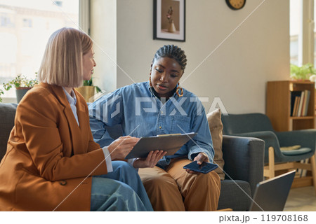 Portrait of adult African American woman talking to female finance expert explaining tax forms and showing clipboard with documents 119708168