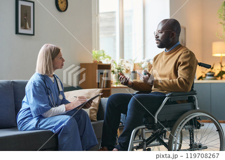 Side view portrait of female doctor talking to African American man with disability during at home medical and rehabilitation visit copy space Side view portrait of female doctor talking to African American man with disability during at home medical and rehabilitation visit copy space 119708267