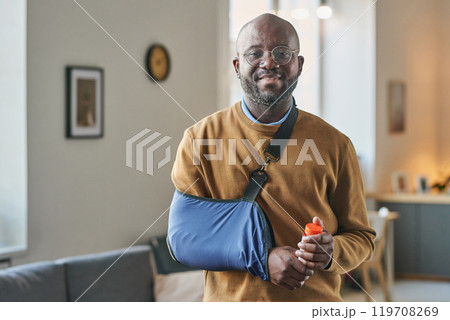 Waist up portrait of smiling adult African American man with broken arm in sling looking at camera and holding bottle of pills copy space 119708269