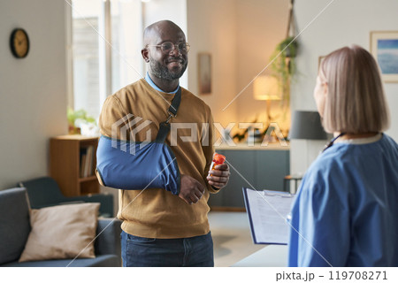 Waist up portrait of smiling adult African American man with broken arm talking to nurse after successful at home med visit copy space 119708271