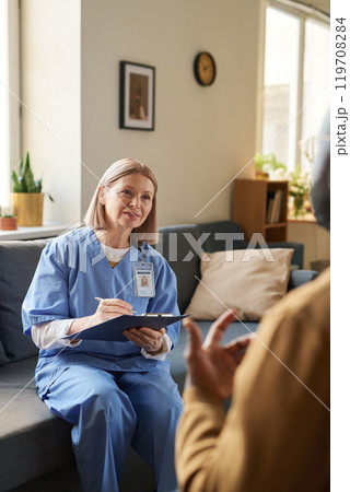 Vertical portrait of smiling female doctor holding clipboard and listening to patient during at home visit Vertical portrait of smiling female doctor holding clipboard and listening to patient during at home visit 119708284