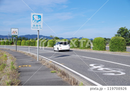 白色のステーションワゴン車が鳥取県境港市の米子鬼太郎空港沿いのメロディー道路を走る光景 119708926