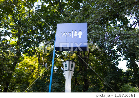 Toilet sign against the background of green trees in the park. White wc sign on blue metal plate 119710561