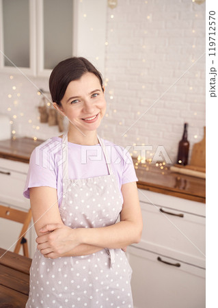 Smiling woman in polka dot apron standing in modern kitchen with warm ambient 119712570