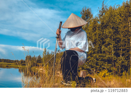 Meditating warrior with katana by the river. Cosplayer in conical hat and mask kneels by the river holding katana, reenacting the scene of meditation and reflection in natural environment. 119712883
