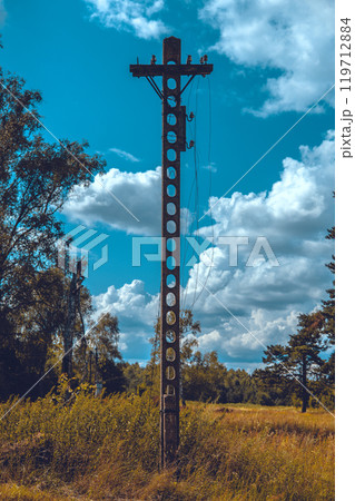 An old concrete power pole with round holes standing in a field with tall grass and trees under a blue sky with white clouds. An old power pole with broken wires. An old concrete power pole with round holes standing in a field with tall grass and trees under a blue sky with white clouds. An old power pole with broken wires. 119712884